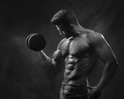 Powerful black and white portrait of a shirtless bodybuilder lifting dumbbells in the gym.