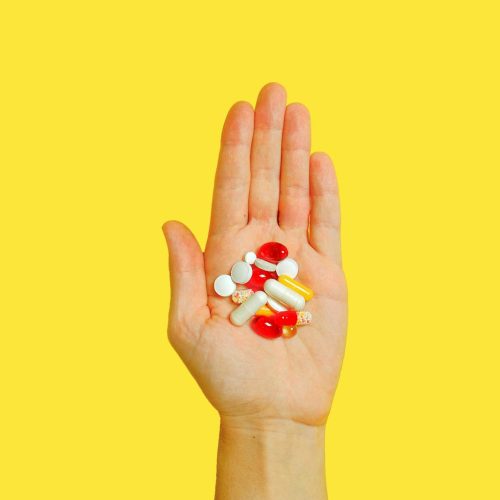 Close-up of an adult hand holding different types of pills against a vivid yellow background.