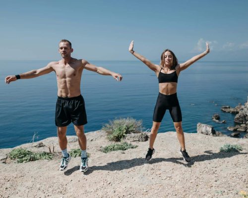 A man and woman perform jumping jacks on a cliff by the ocean under a clear sky.