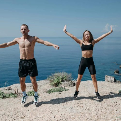 A man and woman perform jumping jacks on a cliff by the ocean under a clear sky.